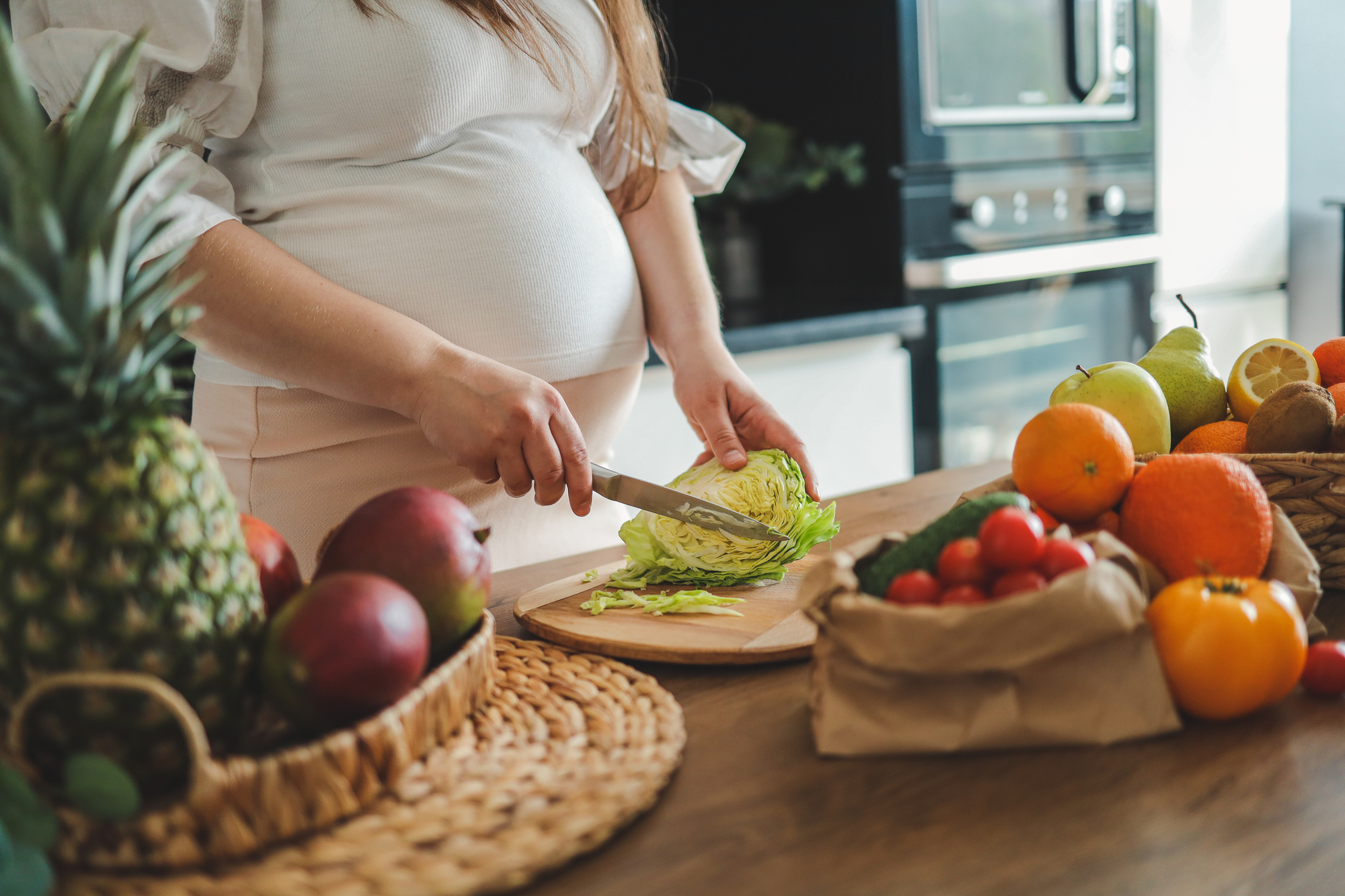 A pregnant woman is seen from the neck down, creating a salad at a counter full of fresh food
