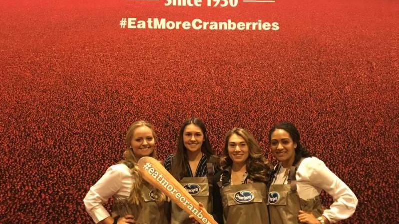 SNA members Valerie Snell, Mackenzie Lombardi, Madison Galascio and Paris Winston in front of a large Ocean Spray poster.