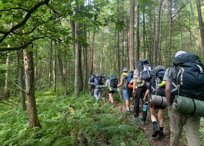 Students wearing outdoor gear and carring backpacks and walking sticks, hike along a trail in the lush green woods.