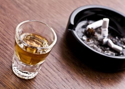A shot glass of whiskey sits next to an ashtray with three discarded cigarettes. 