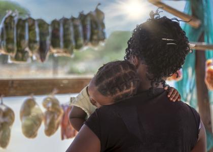 Woman seen from behind holding small child in her arms as she browses for food in an African market