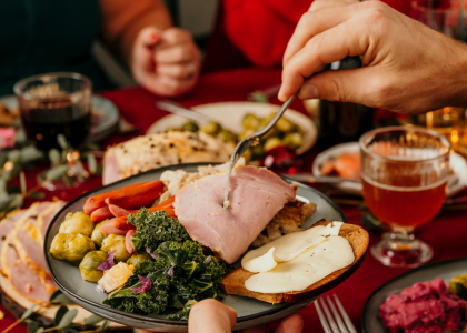 People sitting down for a holiday meal, table filled with a variety of holiday food and drink. Image is focused on a single plate filled with holiday food and a person serving themselves from food on the table. 