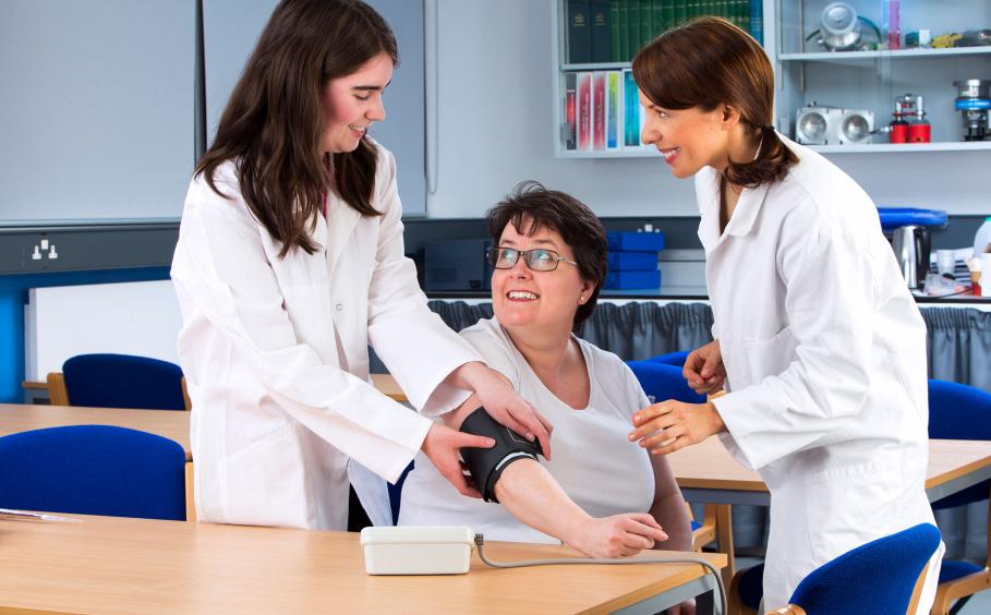 A woman receiving a health check. She is having her blood pressure taken by a medical student who is being supervised by a professional. 