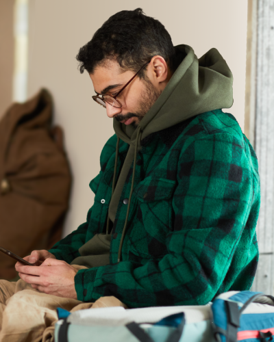 A man wearing a green jacket and hoodie sits on the floor of a shelter with a blanket and holding a smart phone. Others can be seen in the background sitting on the floor as well with bags, blankets, and eating food.