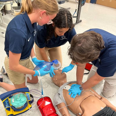 Three athletic training students are set up on the lab floor with a medical manikin practicing CPR techniques. 