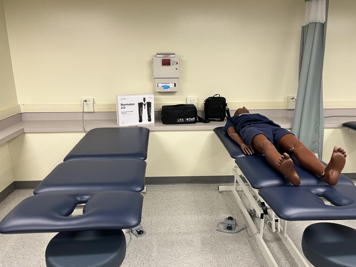 =Two blue treatment tables are pictured, one with a medical manikin laying on the table. Medical equipment is sitting on a counter behind the tables.