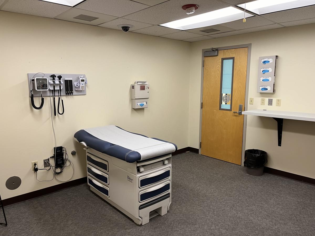 =This exam room has a treatment table, like you would see in a doctor's office, with the wall containing healthcare equipment such as a thermometer and a blood pressure cuff.