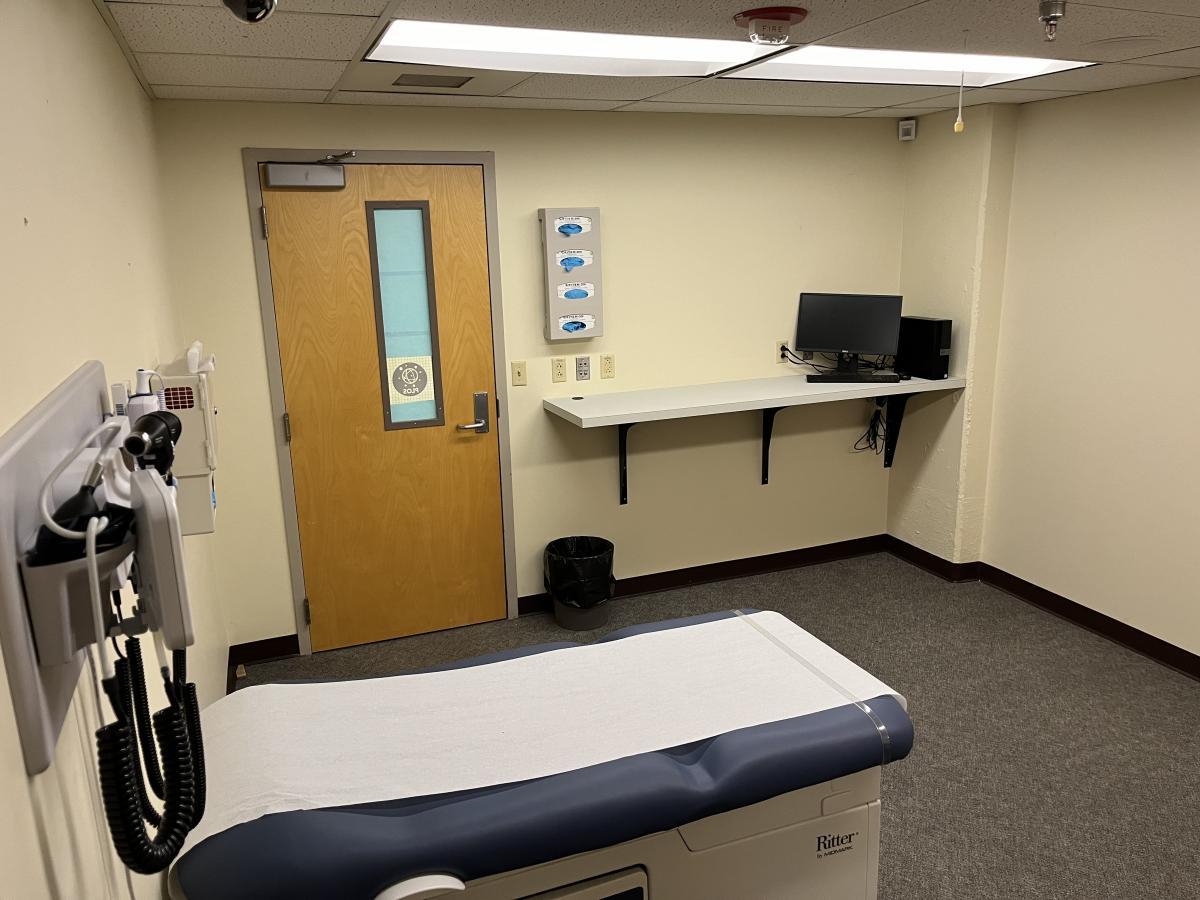 =This is an exam room with a treatment table like you would see in a doctor's office. There is a counter against the wall with computer monitors and a door to the room beside the counter.